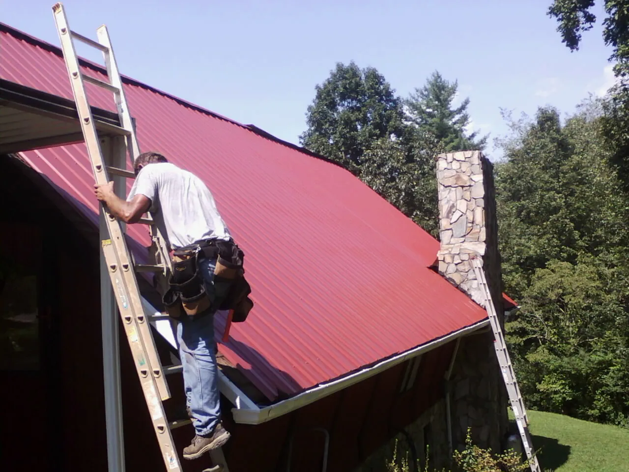 Roofer on a ladder beside a red metal roof and stone chimney — Soffit & Fascia Repair by Roofing Brothers in Betterton